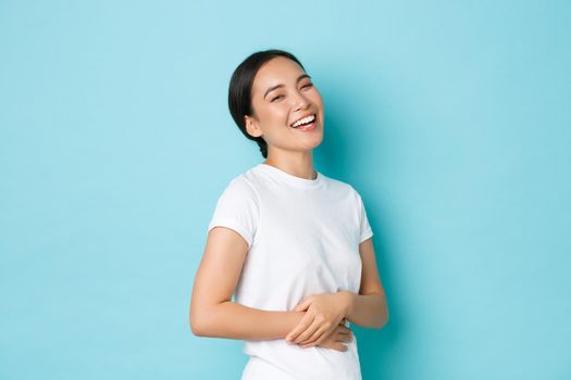 Portrait of carefree beautiful asian girl laughing and smiling pleased, holding hands on belly, standing in determined and confident pose over blue background. Copy space