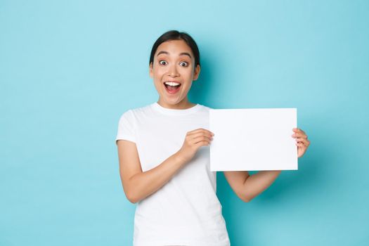 Amazed and excited asian girl in white t-shirt, making announcement, gasping surprised while showing sign, piece of blank paper with your advertisement, standing light blue background.