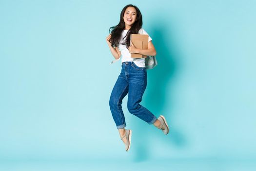 Fashion, back to school and lifestyle concept. Cheerful young asian girl, korean student looking upbeat, holding backpack and notebooks, jumping upbeat over light blue background.