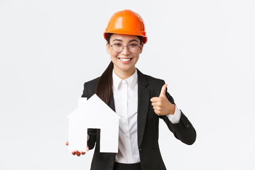 Portrait of young asian female construction engineer, architect in safety helmet and business suit holding home maket and show thumbs-up, promoting company service of building houses.