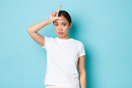 Sad and unconfident in herself asian girl wearing white casual t-shirt, sulking and looking glomy over lost competition, showing loser gesture on forehead, standing blue background.