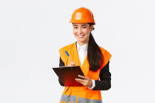Smiling satisfied asian female construction engineer leading inspection at enterprise, wearing safety helmet and reflective jacket, writing down notes and looking pleased result, white background.