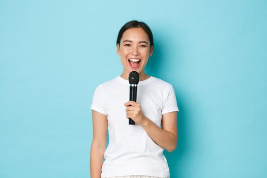 Lifestyle, people and leisure concept. Happy carefree asian girl enjoying performing song, holding karaoke microphone and smiling, singing favorite song, standing blue background.