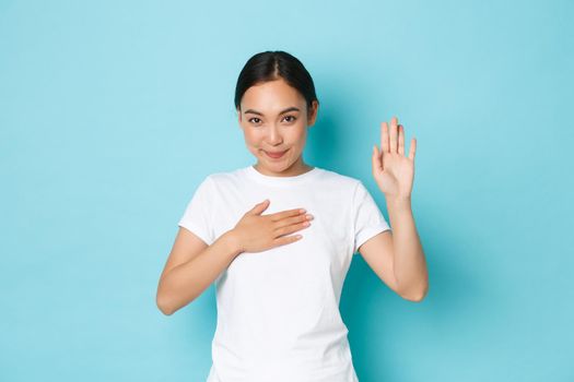 Cute smiling asian girl swearing to tell only truth, making oath or pledge on something. Adorable korean female student hold hand on heart and arm raised while being honest, blue background.