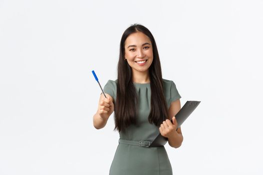 Smiling beautiful asian female shop assistant, employee asking to sign blank or shipping form, giving you pen as holding clipboard with documents, standing white background.