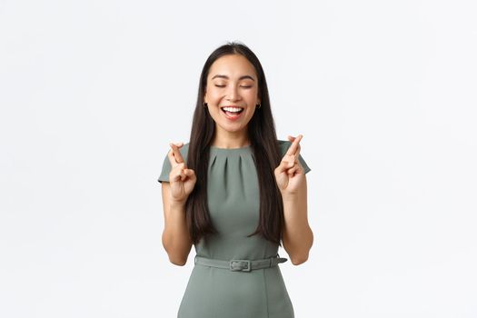 Small business owners, women entrepreneurs concept. Excited happy and hopeful cute asian female making wish, cross fingers good luck close eyes and smiling optimistic, standing white background.