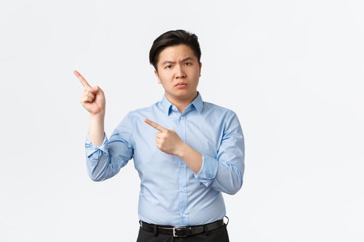 Business, finance and people concept. Disappointed frowning asian businessman in blue shirt pointing upper left corner frustrated, scolding employees, standing white background displeased.