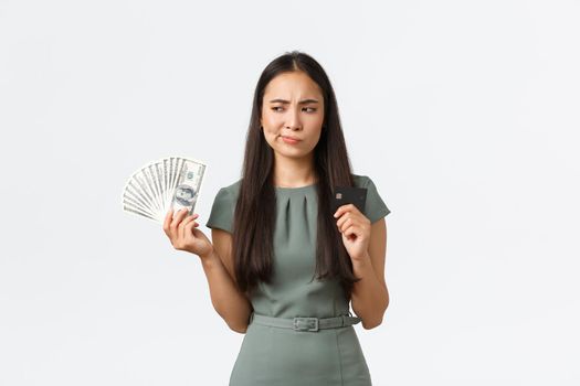 Small business owners, women entrepreneurs concept. Indecisive asian woman thinking while holding credit card and cash, perplexed how to pay for purchase, standing white background thoughtful.