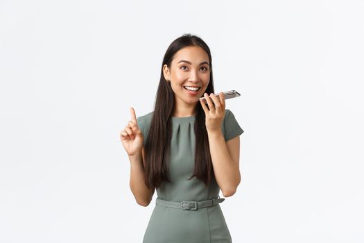 Small business owners, women entrepreneurs concept. Smiling happy asian woman in dress record voice message, talking to personal assistant in phone, looking upbeat, holding smartphone.