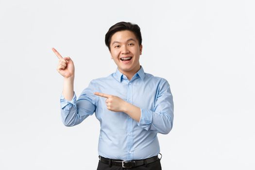 Business, finance and people concept. Enthusiastic smiling asian male entrepreneur in blue shirt, with teeth braces, pointing fingers upper left corner, making announcement, white background.