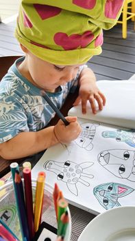 Small toddler girl wearing a funny hat sitting at the table and drawing with pencils