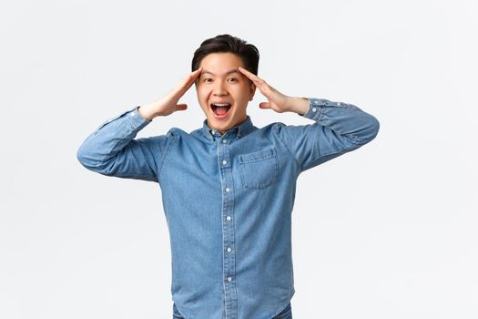 Surprised and happy, relieved asian man rejoicing over great news, holding hands near head and smiling upbeat, got rid of problem, feeling cheerful and upbeat over white background.