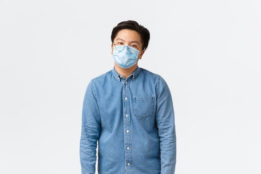 Covid-19, preventing virus, and social distancing at workplace concept. Overworked and tired asian male employee looking reluctant, wearing medical mask, feeling bored, standing white background.