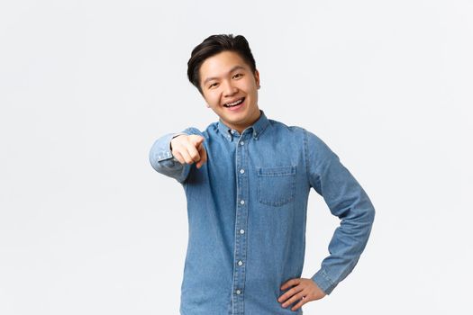 Smiling happy asian handsome guy in blue shirt, have braces, pointing finger at camera, encourage person click link, naming or choosing someone, standing over white background.