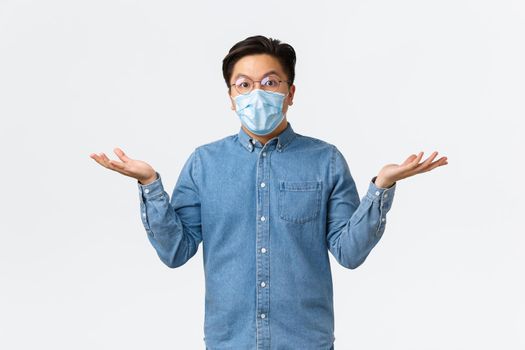 Covid-19, preventing virus, and social distancing at workplace concept. Confused and surprised asian male entrepreneur spread hands sideways and shrugging puzzled, wear medical mask and glasses.