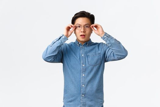 Shocked and impressed asian man put on glasses to see something, looking startled and amazed, witness big event, watching interesting announcement, standing white background.