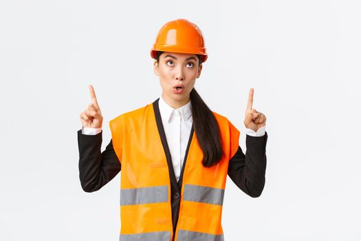 Building, construction and industrial concept. Surprised and curious asian female construction manager, engineer in safety helmet and reflective clothing, pointing fingers up at interesting banner.