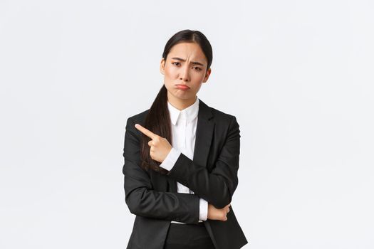 Disappointed gloomy asian female entrepreneur losing, failing job, standing in suit, pouting and pointing finger left at failure. Upset businesswoman sharing bad news, white background.