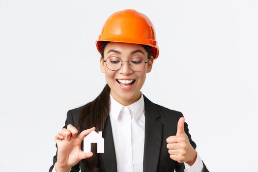 Close-up of happy pleased asian female architect, construction engineer in helmet and suit looking at home miniature and showing thumbs-up, guarantee quality of repairment works, white background.