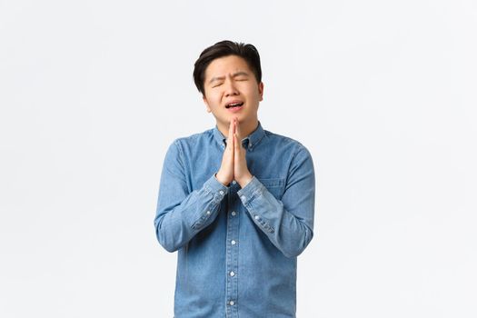 Overworked gloomy and sad asian man begging for help, holding hands together over chest in praying gesture, asking favour, showing remorse, standing white background overwhelmed.