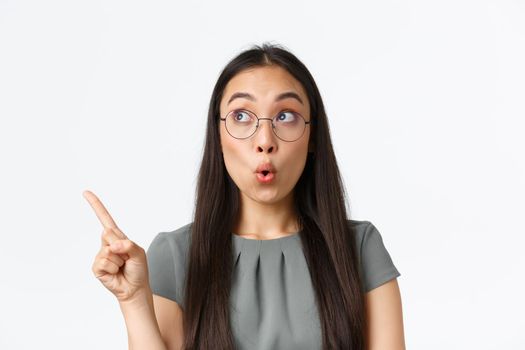 Impressed attractive asian woman checking-out banner with special offer, shop discounts. Female in glasses pointing finger and looking upper left corner with amazed, surprised expression.