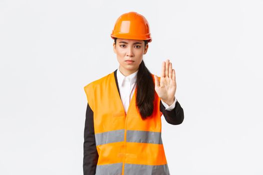 Serious-looking disappointed asian female architect, construction manager at working area wearing safety helmet, showing stop gesture, prohibit action, forbid trespassing, white background.