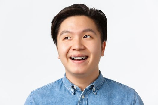 Orthodontics, dental care and stomatology concept. Close-up of hopeful, handsome asian man with teeth braces looking dreamy upper left corner, smiling pleased, standing white background.
