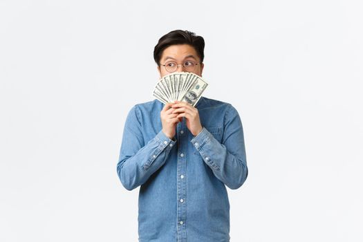 Indecisive young asian man in glasses, looking behind cash, stare away thoughtful, thinking how invest money or what buy, getting ready for shopping, standing white background.