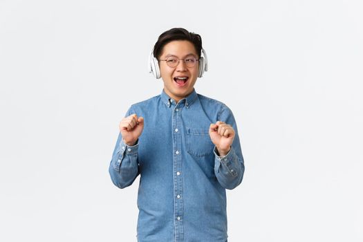 Lifestyle, leisure and technology concept. Carefree smiling funny asian guy vibing and chilling while listening music with wireless headphones, dancing and singing along, white background.