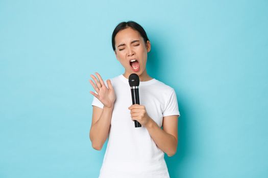 Lifestyle, people and leisure concept. Carefree beautiful asian girl singing sing in microphone with passionate expression, close eyes and gesturing while performing, like karaoke.