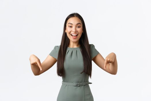 Small business owners, women entrepreneurs concept. Happy beautiful asian businesswoman inviting to check out promo, pointing fingers down at event banner, smiling at client, white background.