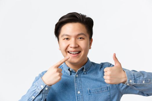 Orthodontics and stomatology concept. Close-up of satisfied asian guy, dental clinic client smiling happy and pointing at his dental braces and showing thumbs-up in approval, recommend.