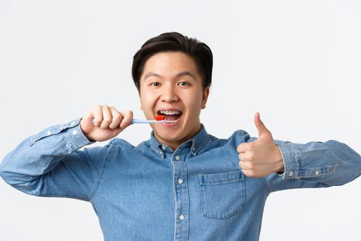 Orthodontics, dental care and hygiene concept. Close-up of satisfied happy asian man brushing teeth with braces, holding toothbrush and showing thumbs-up in approval, white background.