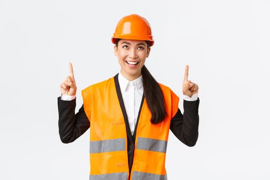 Building, construction and industrial concept. Successful pleased asian female engineer in safety helmet inspect object, looking and pointing fingers up satisfied. Architect pleased with good result.