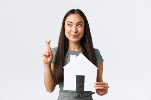 Insurance, loan, real estate and family concept. Hopeful and optimistic smiling asian woman cross fingers and smiling, dreaming of finding new home, holding paper house, making wish.