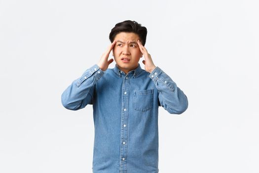 Troubled and uneasy asian man looking perplexed, having complicated situation. Guy with headache touching head and looking away, suffering painful migraine, standing white background.