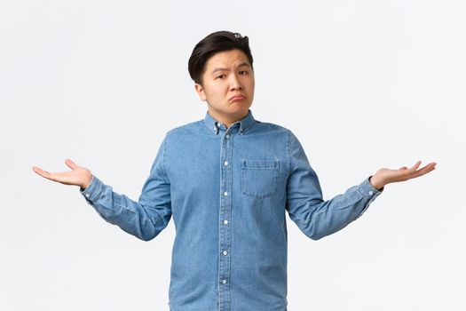 Clueless sad asian young guy dont know anything, spread hands sideways and shrugging unaware, standing clueless, cant understand, standing indecisive over white background without answers.