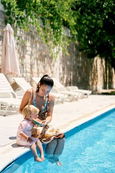 Mom feeds a little girl a watermelon sitting on the side of the pool with turquoise water. High quality photo