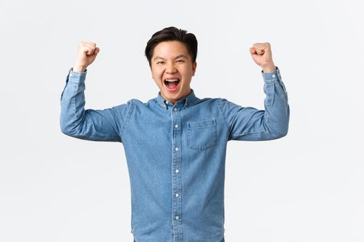 Excited happy asian man with braces feeling success over winning prize, fist pump and shouting yes, rejoicing, triumphing as becoming champion, standing white background celebrating.