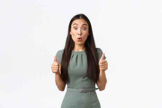 Small business owners, women entrepreneurs concept. Surprised and impressed asian female in dress showing thumbs-up and looking amazed with great work, standing white background.