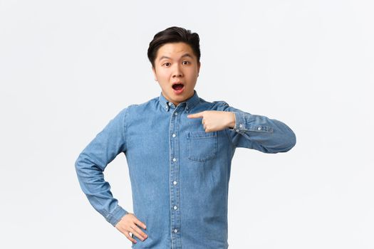 Surprised and questioned asian handsome guy in blue shirt, pointing at himself with curious face, being mentioned or named, chosen from crowd, standing white background.