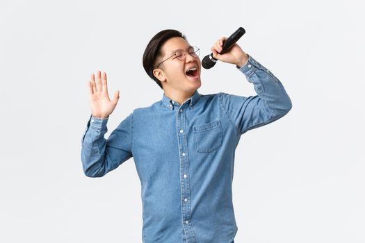 Lifestyle, leisure and people concept. Carefree asian guy in glasses and braces singing song at karaoke bar, holding microphone, performing new single, standing white background joyful.