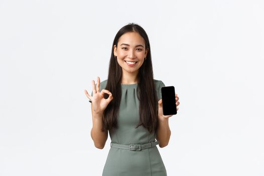 Small business owners, women entrepreneurs concept. Confident smiling asian businesswoman showing new phone application, make okay gesture and introduce mobile app on screen, white background.