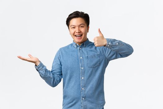 Satisfied handsome asian guy with braces in blue shirt, showing thumbs-up as rating product, raise hand over white background as if holding item, recommend it, demonstrate something.