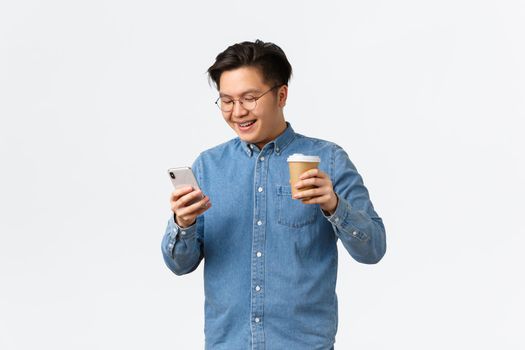 Lifestyle, leisure and technology concept. Smiling asian man, college student having break, drinking takeaway coffee and using mobile phone, texting friend, calling taxi, white background.