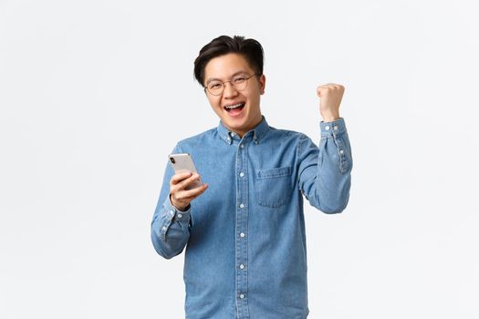 Successful winning asian man in glasses rejocing, fist pump satisfied and smiling as finish level, playing mobile game or got date on dating app, triumphing delighted over white background.