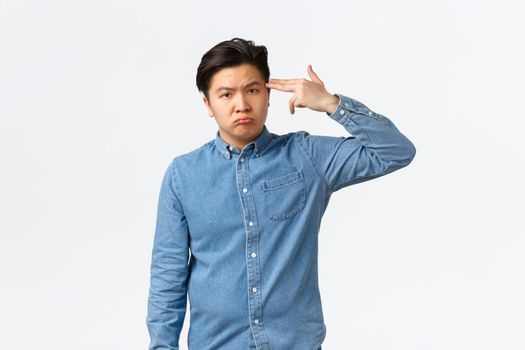 Upset and gloomy asian man feeling tired, overworked guy making gun gesture over head, shooting himself with exhausted sad face, standing white background, wants something end.