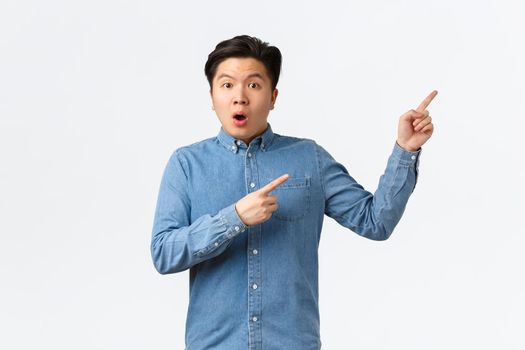 Shocked and astonished asian man asking question about product or announcement. Guy looking startled, pointing fingers upper right corner curious, discussing big event, white background.