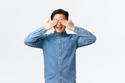 Excited happy asian man with braces, smiling amused as anticipating surprise, holding hands on eyes, standing blindfolded awaiting, standing white background cheerful. Copy space