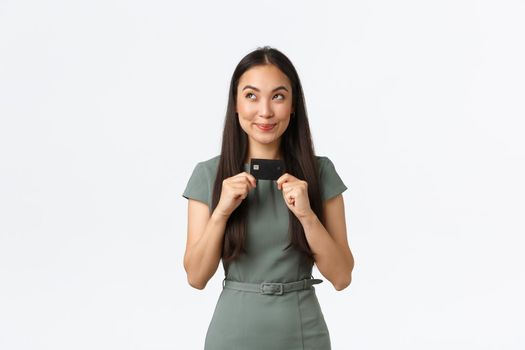 Small business owners, women entrepreneurs concept. Cunning smiling pretty asian woman thinking what buy with credit card, looking away thoughtful and sly, standing white background.